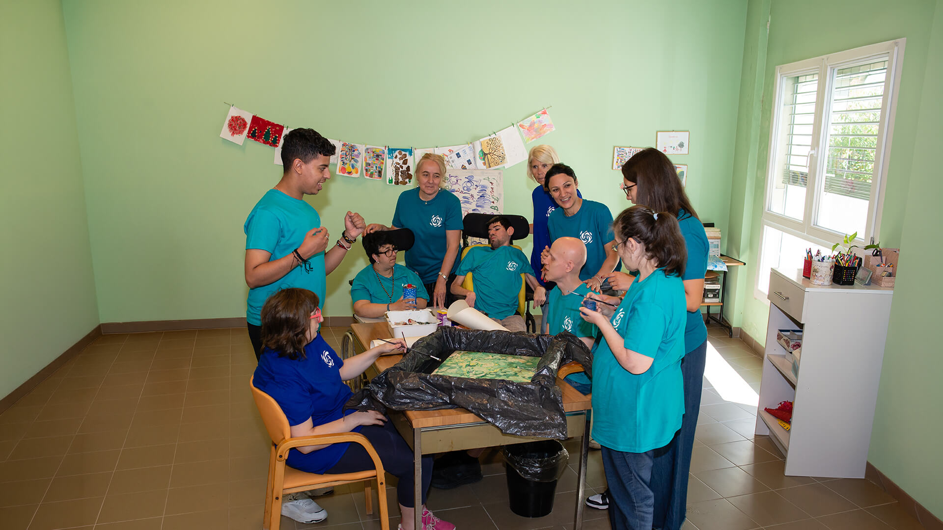 Gruppo sorridente di partecipanti e operatori attorno al tavolo di un laboratorio creativo in una sala luminosa